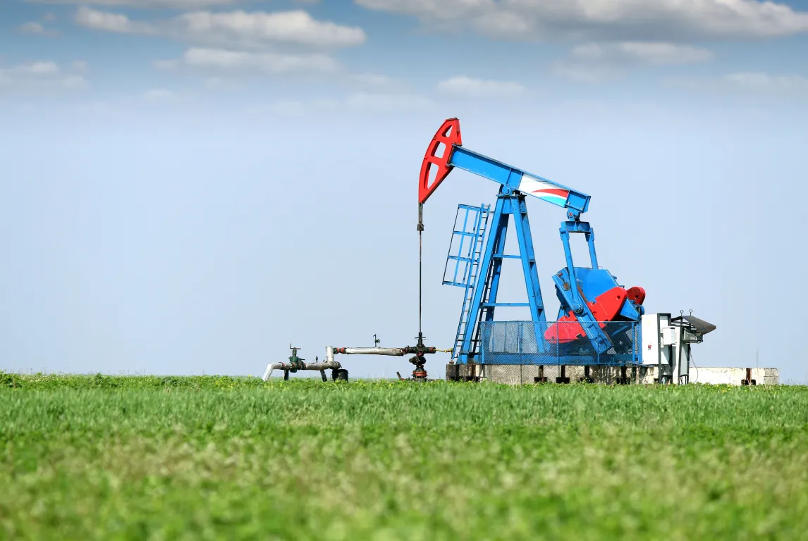 A blue and red oil pumpjack operates in the middle of a green grassy field under a clear blue sky with a few scattered clouds.
