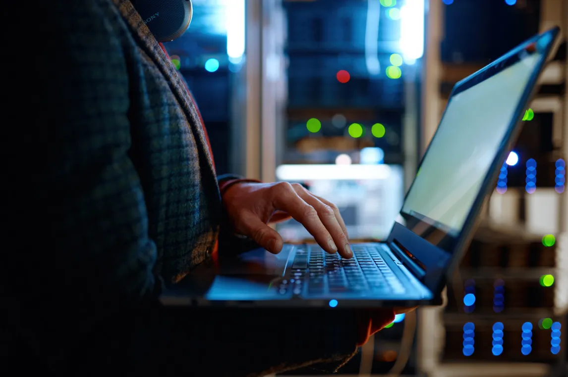 IT technician checking the servers vitals using laptop computer. Male support agent working in a dark network server room