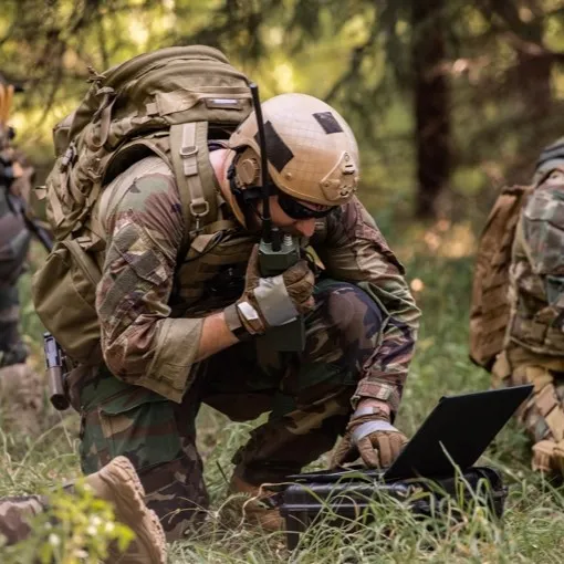 A picture of a soldier holding a radio and looking at a laptop in a field