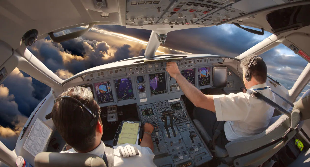 Two pilots wearing headsets operate the controls inside an airplane cockpit, with illuminated flight instruments and dramatic cloud formations visible through the windshield at sunset.