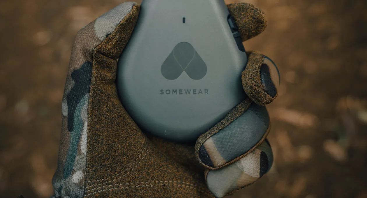 Gloved hand holding a gray Somewear satellite communication device with the Somewear logo, against a blurred outdoor background.