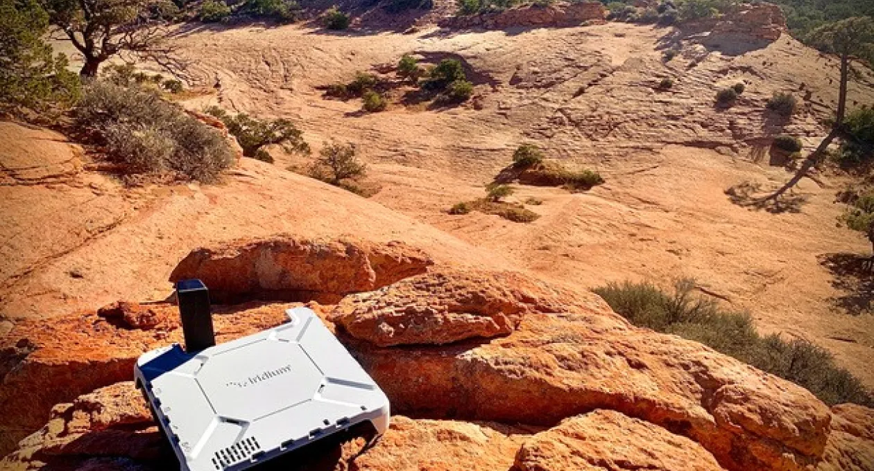 A white Iridium satellite communication device sits on a rocky ledge overlooking a desert canyon landscape with scattered trees and clear blue sky.Mar