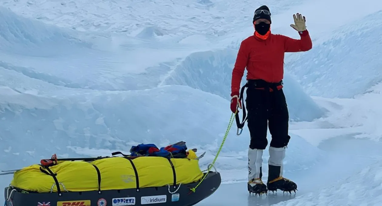 A polar explorer wearing a red jacket, face covering, and crampons stands on icy terrain beside a yellow-loaded sled, waving while surrounded by deep snow and glacial formations.