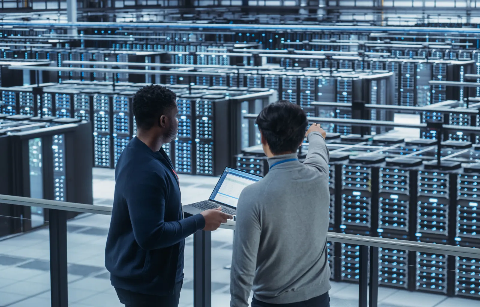 Two Multiethnic Male Data Center System Administrators and IT Specialists Talk, Use Laptop. Information Technology Engineers work on Cyber Security Network Protection in Cloud Computing Server Farm.