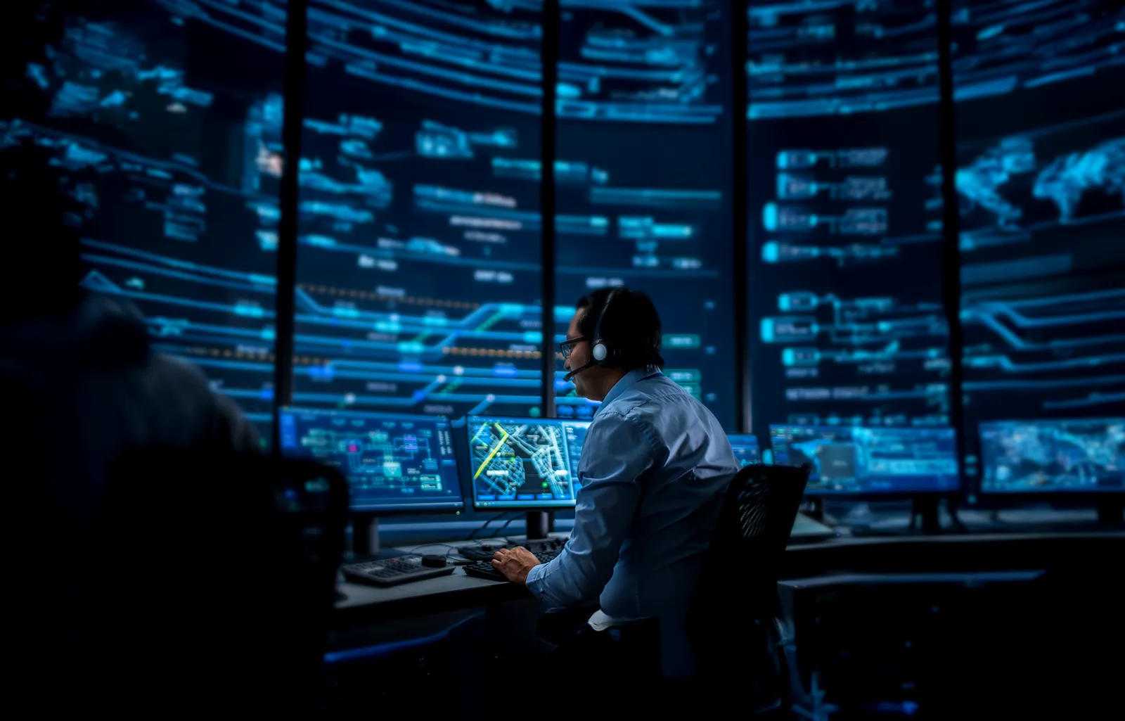 Professional IT Technical Support Specialist and Software Developer Working on Computer in Monitoring Control Room with Digital Screens. Employee Uses Headphones with Mic and Talking on a Call.