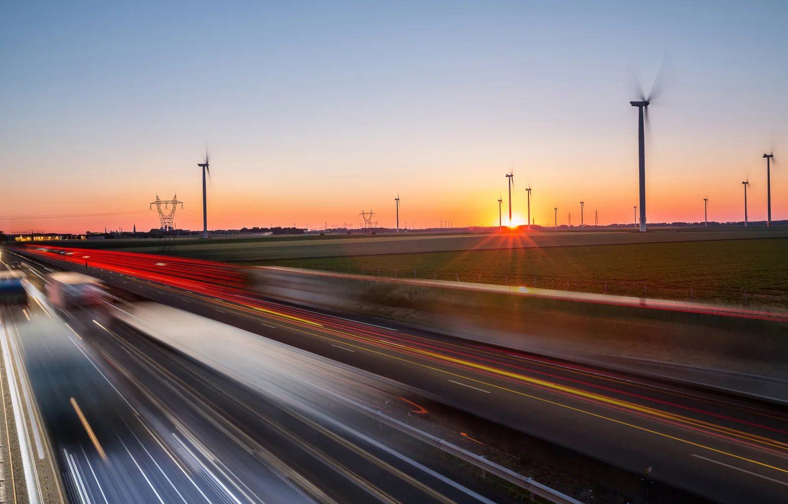A highway at sunset with streaks of red and white lights from passing vehicles, wind turbines spinning in the background, and the sun setting over open fields, symbolizing clean energy and transportation.
