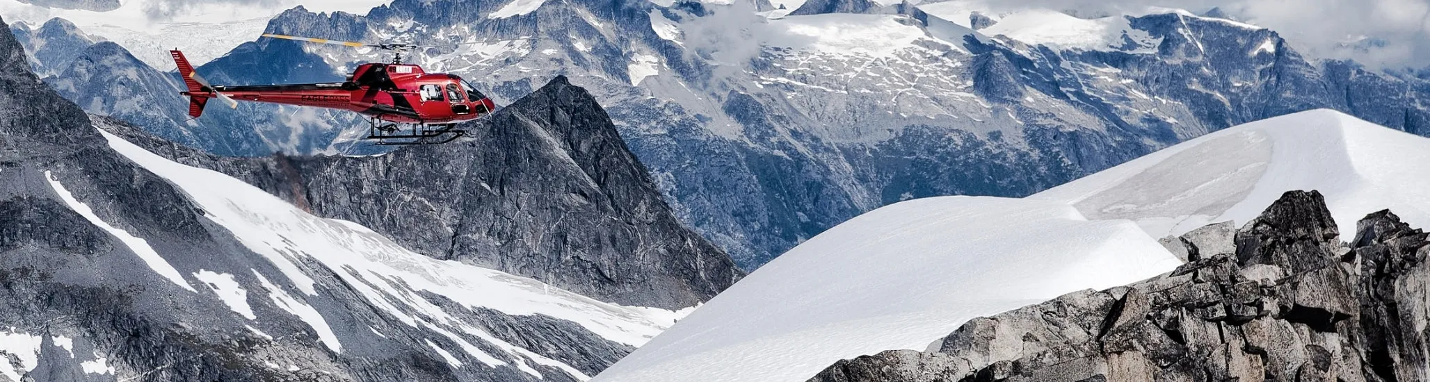A red helicopter flies over rugged snow-covered mountains and rocky peaks, with distant glaciers and clouds under a bright blue sky.