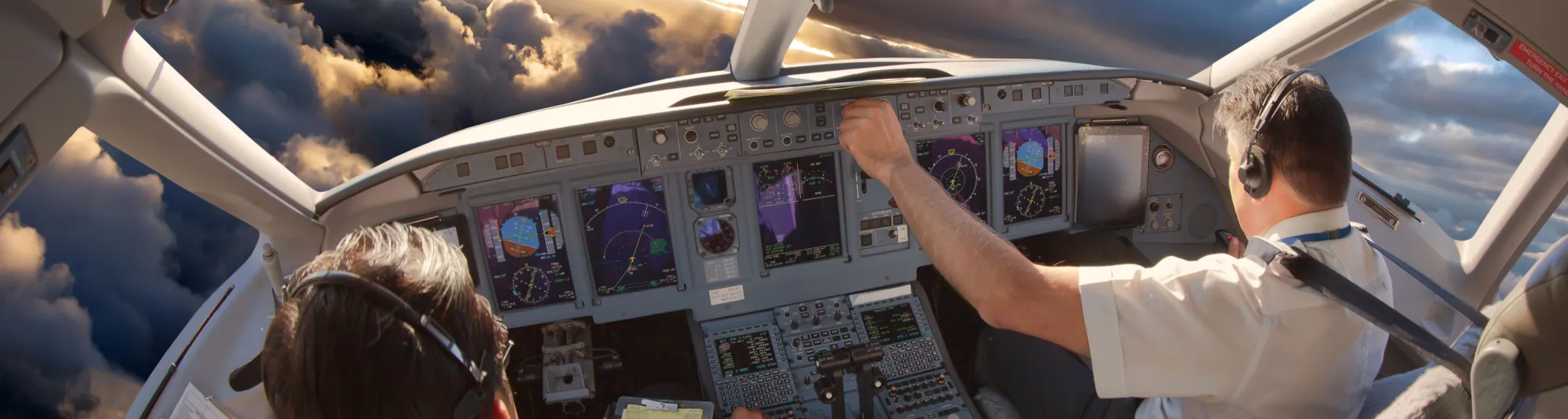 Two pilots wearing headsets operate the controls inside an airplane cockpit, with illuminated flight instruments and dramatic cloud formations visible through the windshield at sunset.