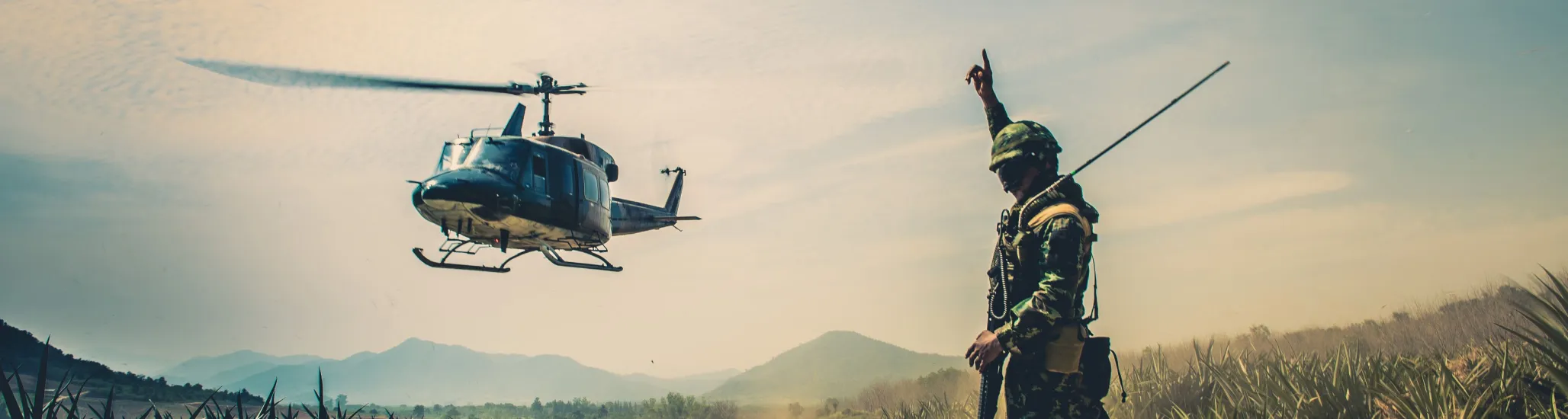 A soldier stands on a dirt road signaling to a helicopter hovering nearby, surrounded by fields and distant mountains under a bright blue sky. Dust swirls from the rotor blades as the aircraft prepares to land or take off.