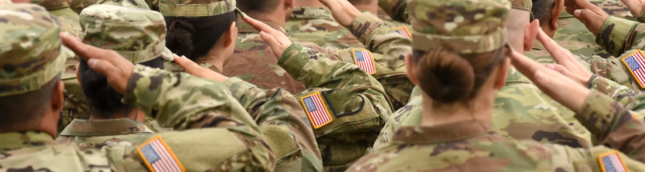 A group of U.S. soldiers in camouflage uniforms stand in formation, saluting with their right hands raised. The American flag patches on their sleeves are visible as they face forward in unison