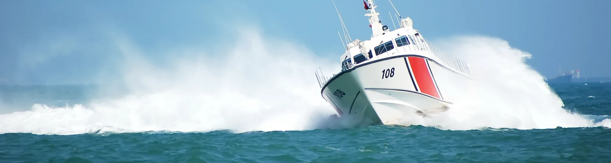 A white and red coast guard vessel numbered “108” speeds through rough blue waters, cutting through waves and creating a large spray under a clear blue sky.