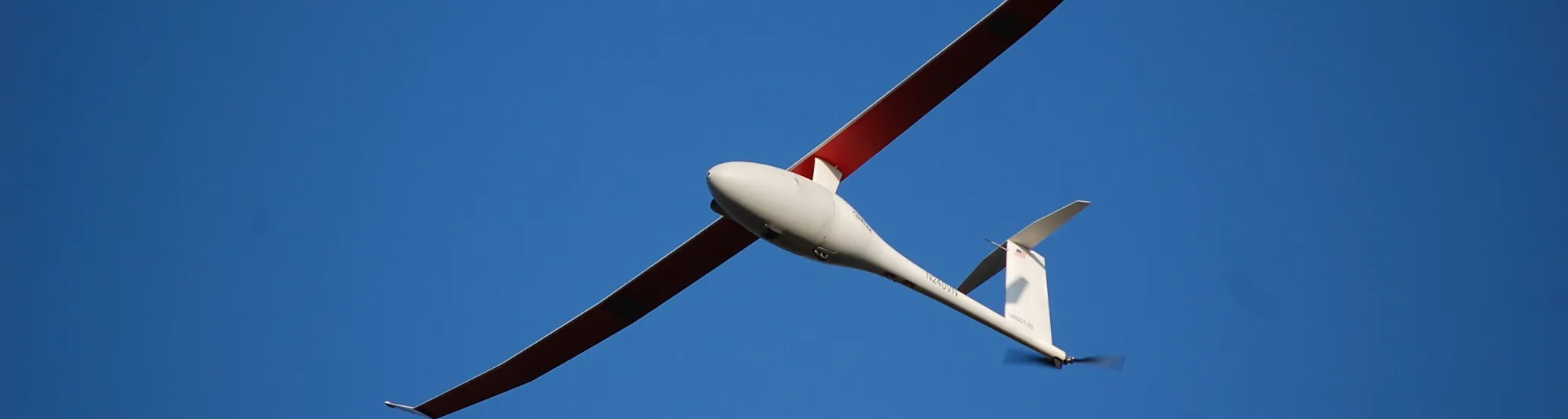 A sleek, long-winged unmanned aircraft with red and white wings soars through a clear blue sky.