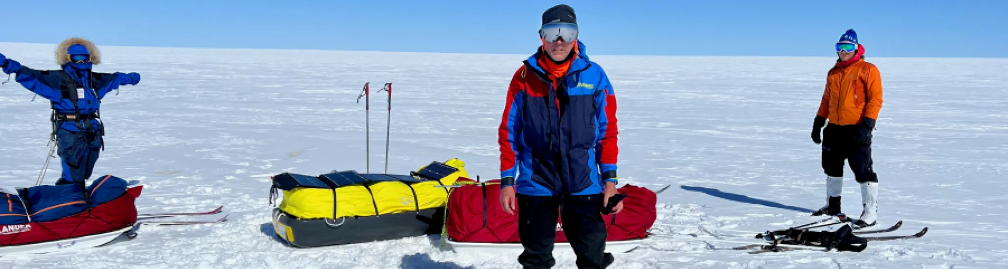 Three expedition members stand on a snowy, wide-open polar landscape with sleds loaded with bright yellow gear and solar panels. The group is dressed in heavy cold-weather clothing and ski equipment under a clear blue sky.