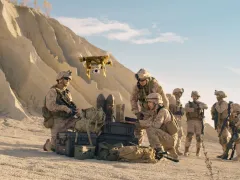 A group of soldiers in desert camouflage operate a small drone in a rocky, arid landscape. Some kneel around equipment cases and laptops while others stand watch, with rifles and tactical gear visible under clear skies and bright sunlight.
