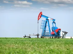 A blue and red oil pumpjack operates in the middle of a green grassy field under a clear blue sky with a few scattered clouds.