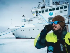 Person in winter clothing holding a device and standing next to a large vessel.