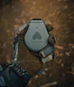 Gloved hand holding a gray Somewear satellite communication device with the Somewear logo, against a blurred outdoor background.