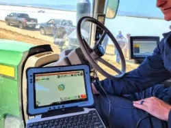 A person in a tractor cab uses a rugged tablet showing a GPS field map, with vehicles and people visible outside.