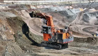 A yellow front-end loader moves piles of black coal in an industrial mining area, with tire tracks visible on the ground and storage structures in the background.