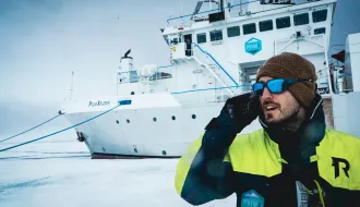 Person in winter clothing holding a device and standing next to a large vessel.
