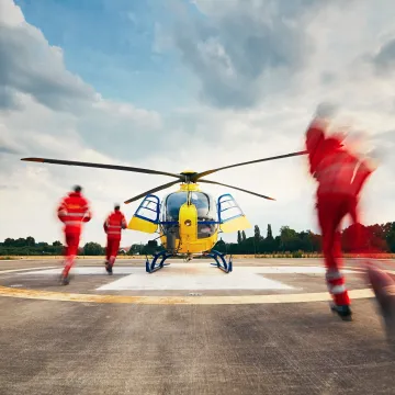 A yellow and blue rescue helicopter sits on a helipad as three paramedics in red uniforms run toward it, ready for takeoff under a cloudy evening sky.