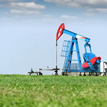 A blue and red oil pumpjack operates in the middle of a green grassy field under a clear blue sky with a few scattered clouds.