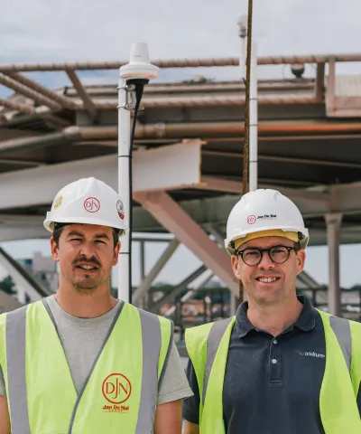 Two men stand on a ship in hard hats and safety vests