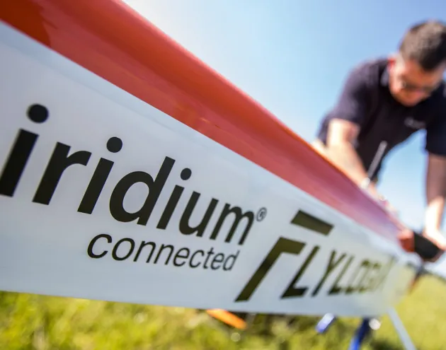 Close-up of an aircraft fuselage labeled “Iridium connected Flylogix,” with a person in the background working on the plane outdoors under a clear blue sky.