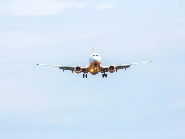 An airplane moves closer to the runway, surrounded by a light gray sky. It angles downward, preparing for a smooth landing at the busy airport, showcasing its landing gear and bright lights.