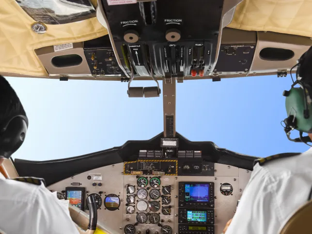 Two pilots wearing headsets sit in the cockpit of an aircraft, surrounded by flight instruments and controls, with a clear blue sky visible through the windshield.