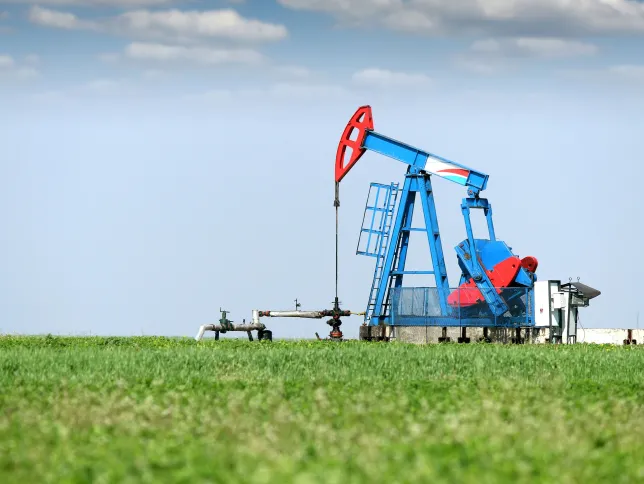 A blue and red oil pumpjack operates in the middle of a green grassy field under a clear blue sky with a few scattered clouds.