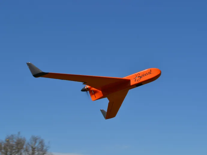 An orange Boreal UAV (unmanned aerial vehicle) with black-tipped wings flies through a clear blue sky, captured in midair during flight.