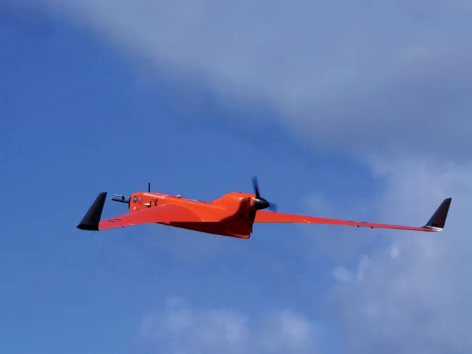 An orange fixed-wing drone with black wingtips and a front propeller flies through a partly cloudy blue sky.