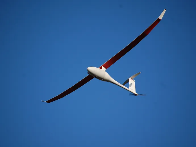 A sleek, long-winged unmanned aircraft with red and white wings soars through a clear blue sky.