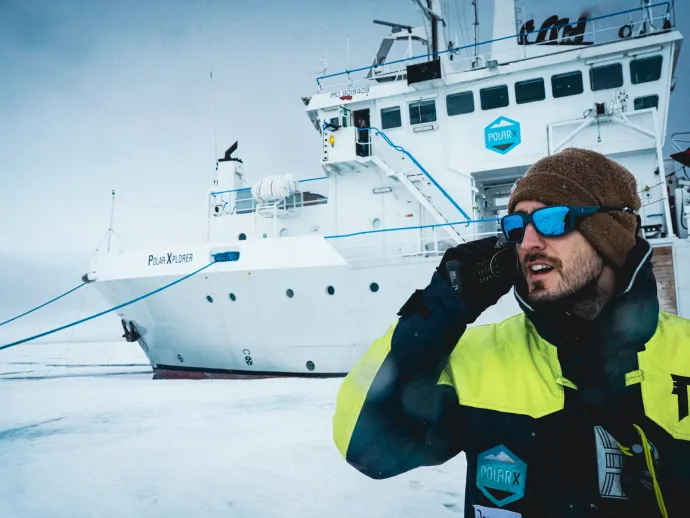 Person in winter clothing holding a device and standing next to a large vessel.