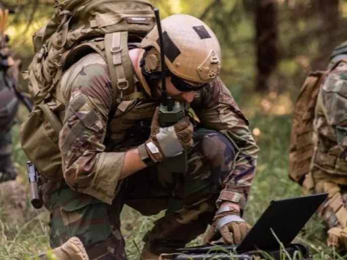 A picture of a soldier holding a radio and looking at a laptop in a field