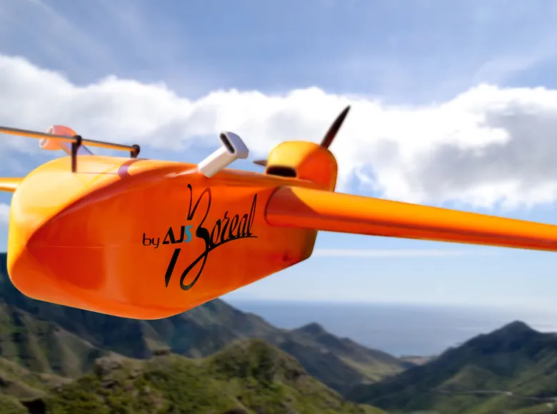 An orange Boreal UAV (unmanned aerial vehicle) with sensors and a propeller flies above green mountain peaks under a partly cloudy sky, with the ocean visible in the distance.