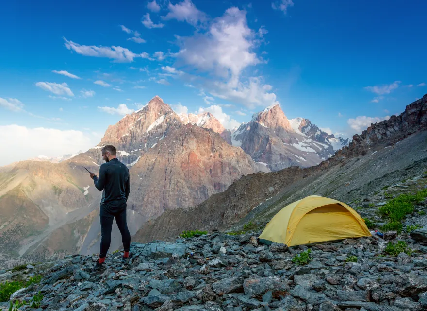 Silhouette of man in wild mountain landscape walk along yellow camping tent holding radio transmitter connection with team blue sky sunny evening