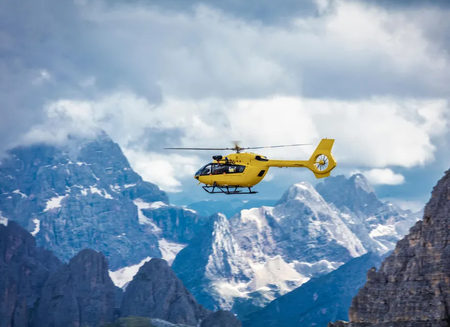 A bright yellow rescue helicopter flies over a rugged mountain range with dramatic rocky peaks and patches of snow beneath a cloudy sky.