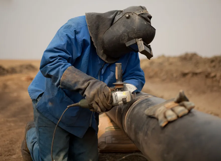 A worker wearing a blue jacket, heavy gloves, and a protective welding helmet kneels on the ground while grinding or welding a large metal pipeline in a dirt construction area.