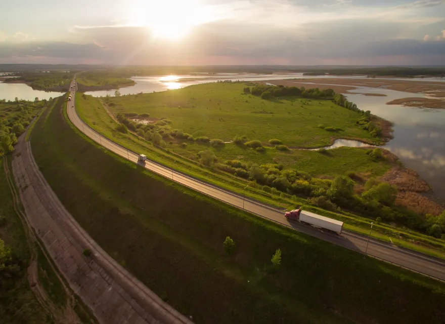 A red semi-truck driving along a winding highway beside green fields and water at sunset, with golden light reflecting off the landscape and distant horizon.