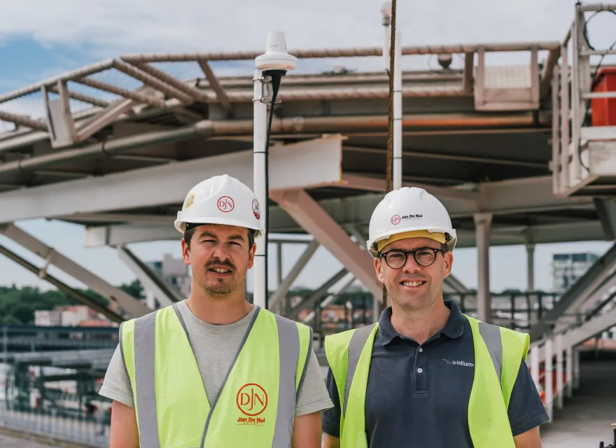 Two men stand on a ship in hard hats and safety vests