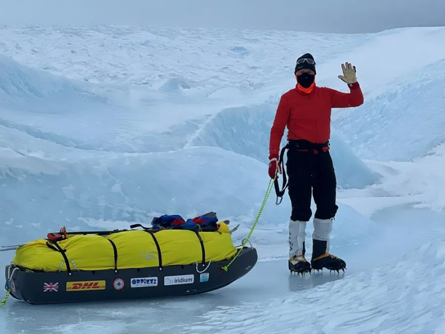 A polar explorer wearing a red jacket, face covering, and crampons stands on icy terrain beside a yellow-loaded sled, waving while surrounded by deep snow and glacial formations.
