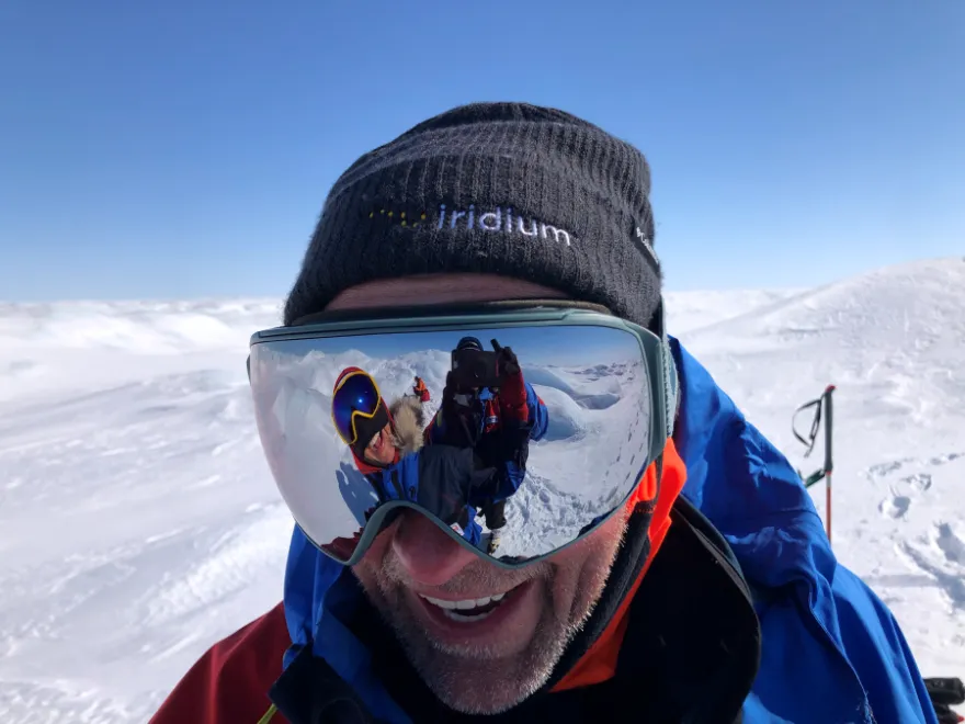 A bundled-up polar expedition member smiles while wearing reflective ski goggles and an Iridium-branded beanie. The snowy landscape is visible behind them, and the goggles show a reflection of another team member taking the photo on the icy terrain.