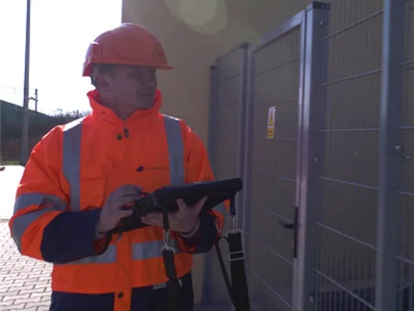 A worker in a hard hat and high-visibility jacket uses a tablet while standing beside a metal fence.