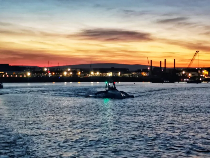 The Mayflower Autonomous Ship (MAS) travels across a harbor at dusk.