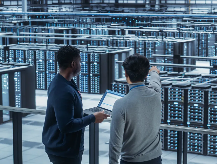 Two Multiethnic Male Data Center System Administrators and IT Specialists Talk, Use Laptop. Information Technology Engineers work on Cyber Security Network Protection in Cloud Computing Server Farm.
