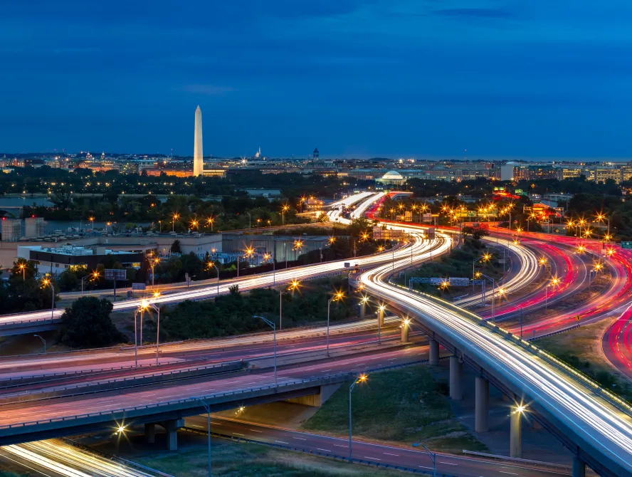 Washington D.C. cityscape at dusk with rush hour traffic trails on I-395 highway. Washington Monument, illuminated, dominates the skyline.