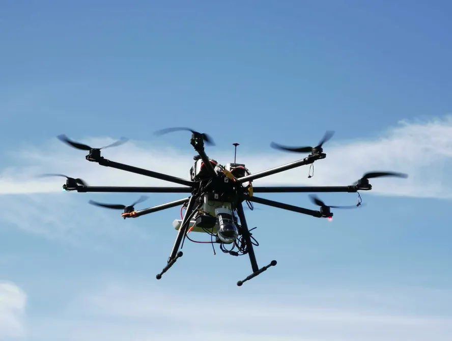 A large black multi-rotor drone equipped with cameras and sensors hovers in the sky against a backdrop of light clouds and blue sky.