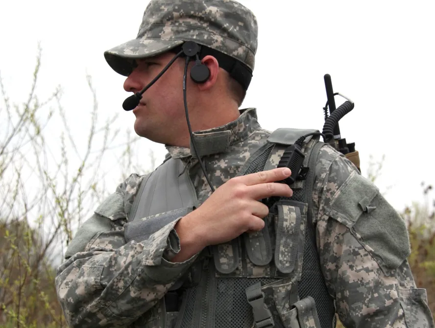 A soldier wearing camouflage uniform and tactical gear uses a handheld radio while speaking into a headset microphone during field communication.